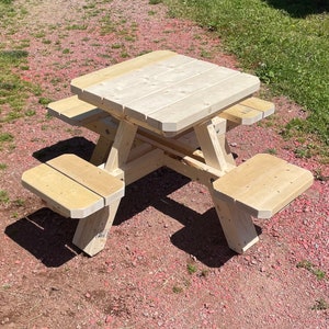Puede incluir: Pequeña mesa de picnic de madera sin terminar con bancos adjuntos. La mesa tiene una parte superior cuadrada y está construida con madera de color claro. Los bancos están unidos al marco de la mesa, creando un conjunto de asientos al aire libre compacto.