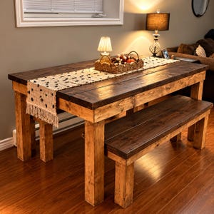 May include: A rectangular wooden dining table with a dark brown stained top and natural light brown legs, accompanied by a matching wooden bench. A cream-colored woven table runner with black tufted accents and fringe sits on the table surface.