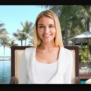 May include: A woman smiles while sitting in a chair in front of a tropical resort pool. Palm trees and a white umbrella are visible in the background.