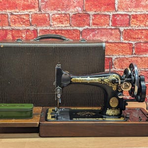 May include: A vintage black Singer sewing machine with intricate gold scrollwork patterns, mounted on a wooden base. It includes a dark brown textured carrying case and a small rectangular olive green metal storage tin placed on the wooden extension.