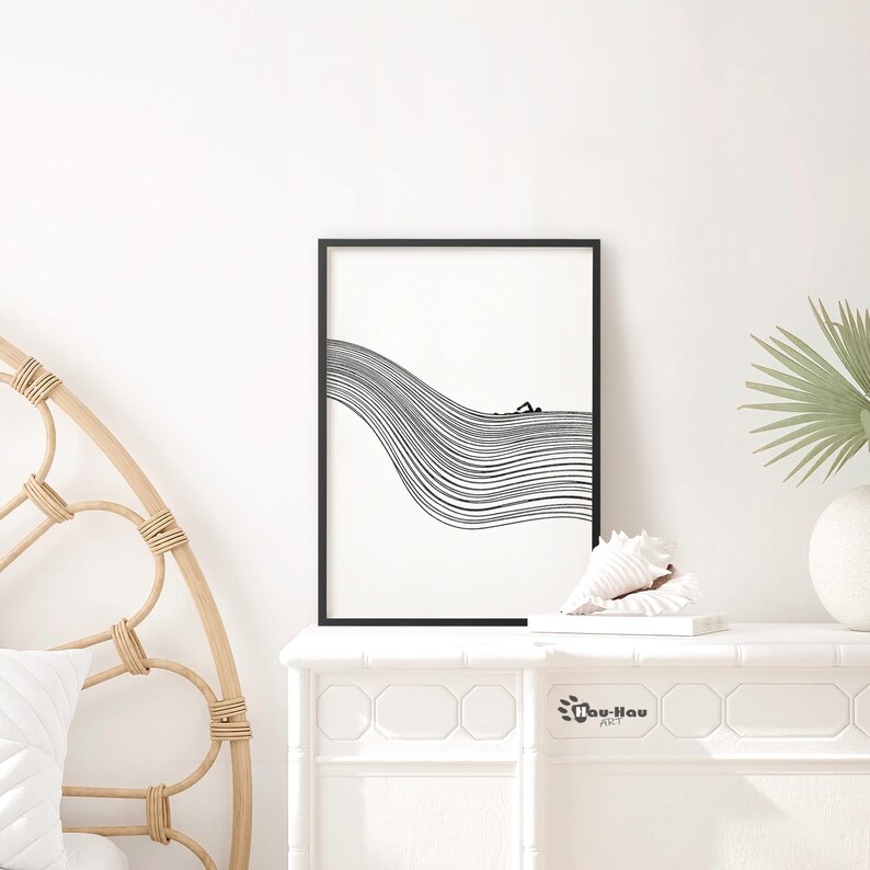 May include: Black and white line art print of a person swimming in a wave. The print is framed in a black frame and is displayed on a white shelf with a seashell and a book.