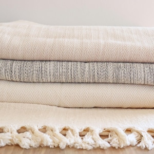 May include: A stack of four white and gray woven throw blankets with fringe edges. The blankets are folded and arranged on a wooden surface.