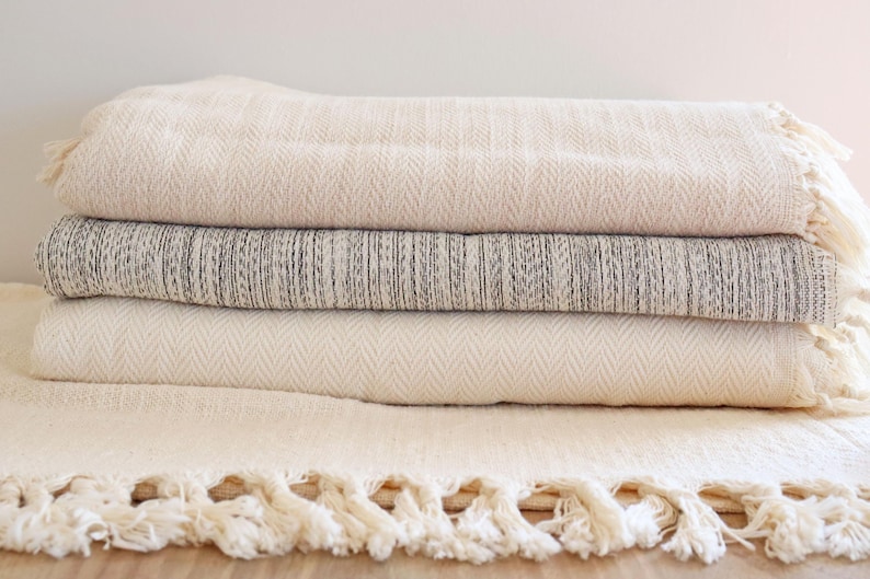 May include: A stack of four white and gray woven throw blankets with fringe edges. The blankets are folded and arranged on a wooden surface.