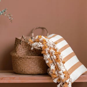 Op de afbeelding: Een geweven mand met een wit en geel gestreept deken met franjes eroverheen gedrapeerd. De mand en het deken staan op een houten plank tegen een bruine muur.