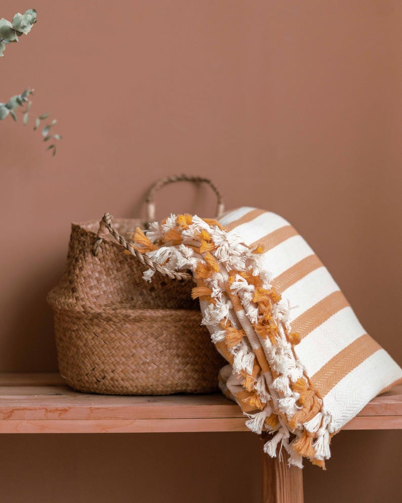 Op de afbeelding: Een geweven mand met een wit en geel gestreept deken met franjes eroverheen gedrapeerd. De mand en het deken staan op een houten plank tegen een bruine muur.