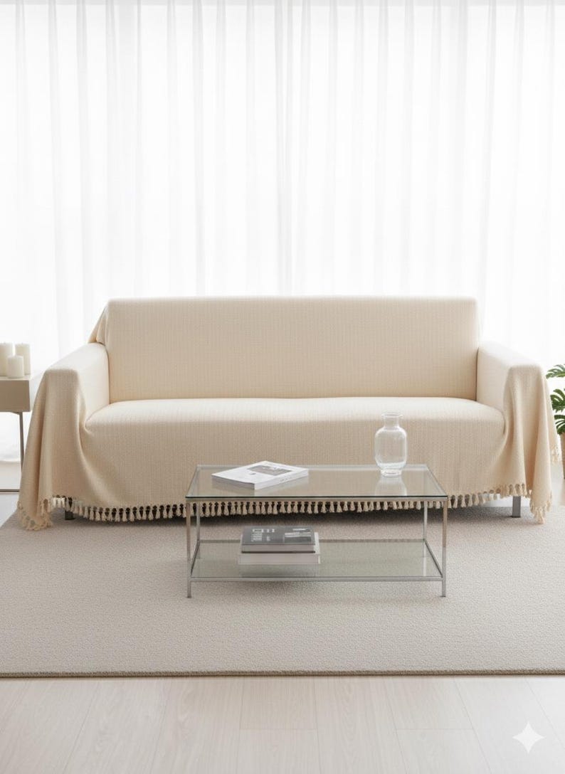 May include: A cream-colored sofa with a matching throw blanket and a glass coffee table. The throw blanket has a fringe trim. A clear glass vase and books are on the table. The room has a neutral color palette.