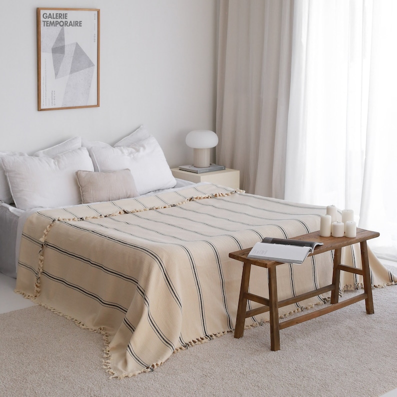 May include: A beige and black striped throw blanket with fringe trim is draped over a bed with white linens. A wooden stool with a book on top sits in front of the bed.