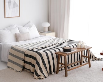 May include: A bed with a cream-colored blanket featuring wide black and gray stripes. The blanket has a fringed edge. A small wooden bench sits at the foot of the bed, and a framed poster hangs on the wall. White pillows and bedding complete the scene.