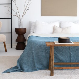 May include: A blue cotton throw blanket draped over a bed with white linens. A wooden bench sits in front of the bed with a black box and a book on top.