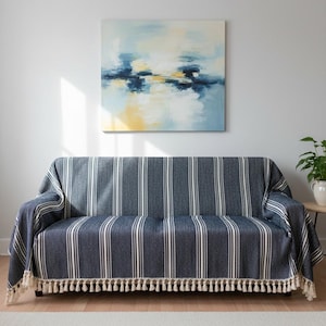 May include: A navy blue and white striped sofa cover with a cream-colored tassel fringe. The cover is draped over a dark brown sofa. An abstract painting with blue, yellow, and white hues hangs on the wall above the sofa.