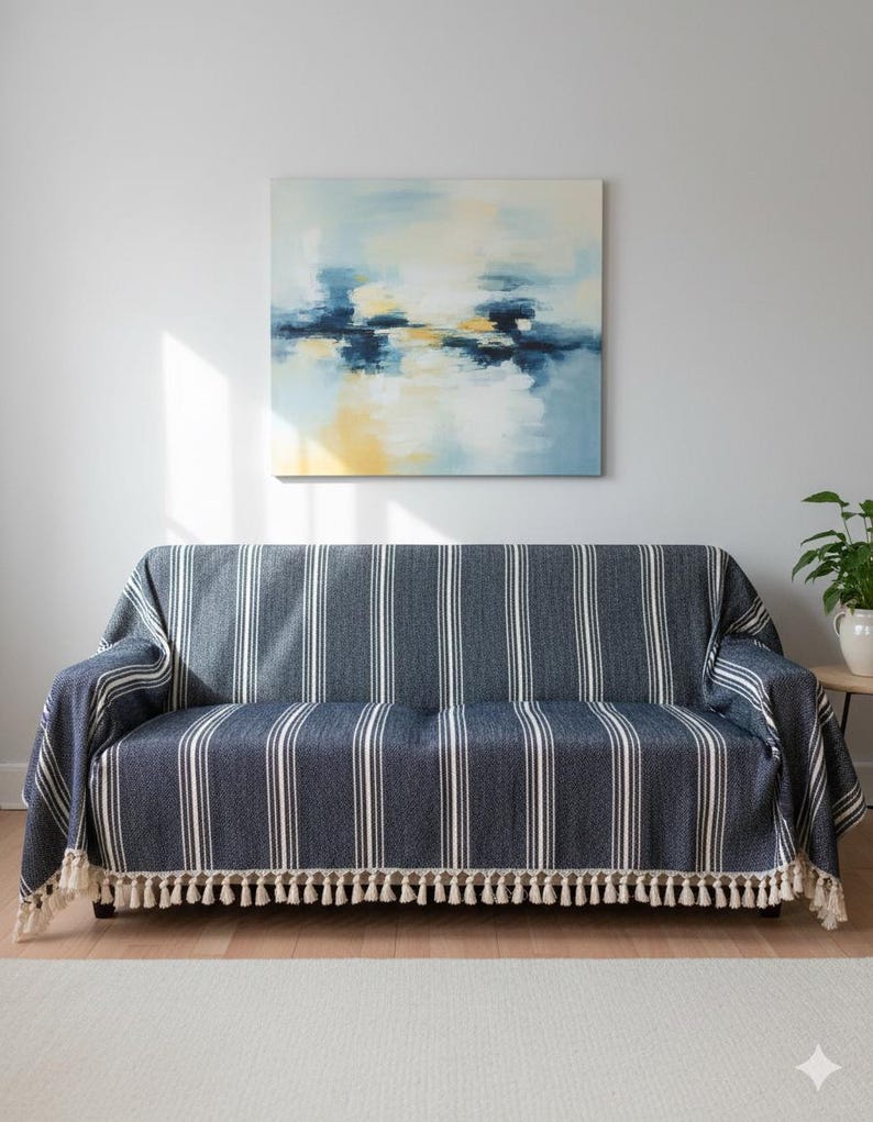 May include: A navy blue and white striped sofa cover with a cream-colored tassel fringe. The cover is draped over a dark brown sofa. An abstract painting with blue, yellow, and white hues hangs on the wall above the sofa.