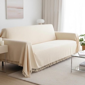 Peut inclure: Une couverture de canapé crème avec un tissage texturé et une frange de pompons sur le bord inférieur. La couverture est drapée sur un canapé beige, créant un aspect chaleureux et invitant. La pièce a une palette de couleurs neutres.