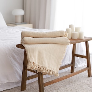 Op de afbeelding: Een cr&egrave;mekleurige deken met franjes aan de randen is gevouwen en geplaatst op een houten kruk. De kruk staat in een slaapkamer met een wit bed en een witte lamp op een nachtkastje.