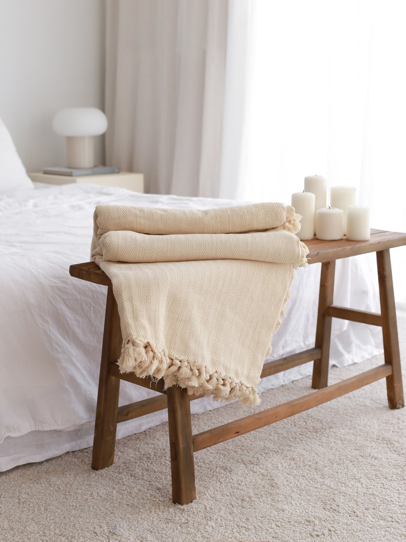 Op de afbeelding: Een cr&egrave;mekleurige deken met franjes aan de randen is gevouwen en geplaatst op een houten kruk. De kruk staat in een slaapkamer met een wit bed en een witte lamp op een nachtkastje.