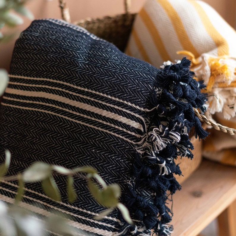May include: A navy blue and beige striped throw blanket with fringe detail. The blanket has a herringbone weave pattern and is folded in a basket with a yellow and white striped blanket.