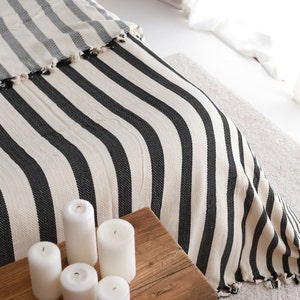 Op de afbeelding: Een wit en zwart gestreept deken met een franje rand. Het deken is over een bed gelegd en er staan witte kaarsen op een houten tafel op de voorgrond.