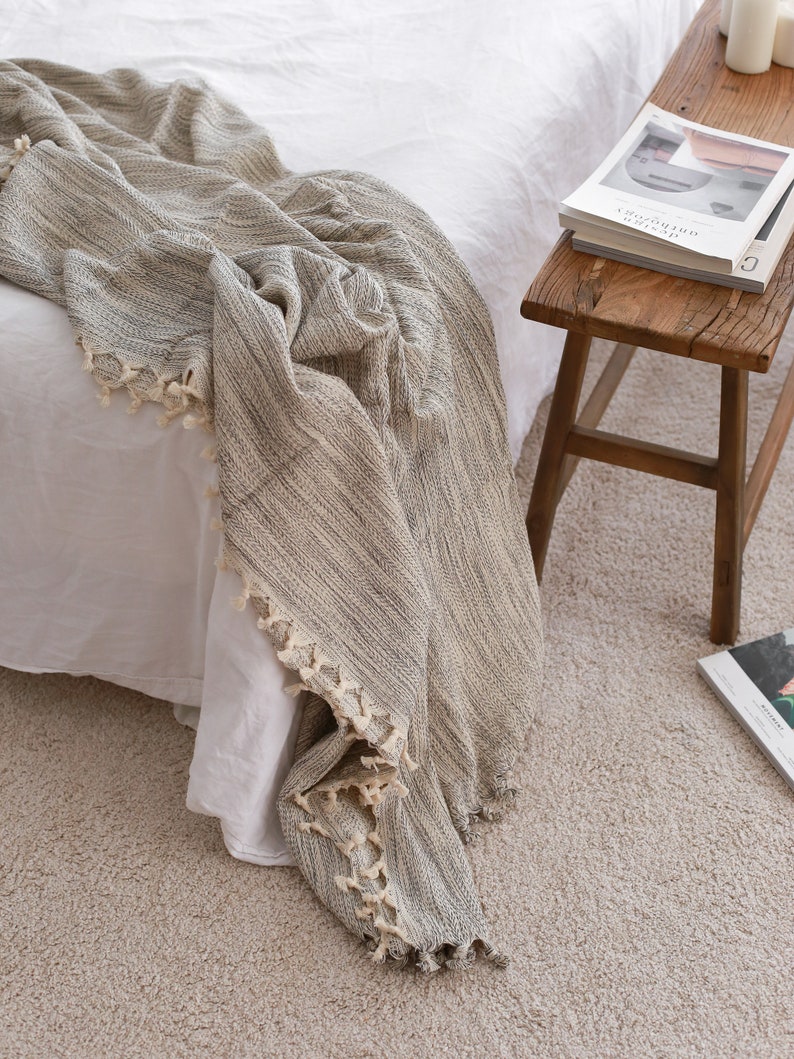 Op de afbeelding: Een grijs en wit gestreepte deken met franjes. De deken is gedrapeerd over een wit bed en een houten kruk met een boek erop.