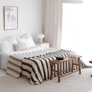 Op de afbeelding: Een zwart-wit gestreepte deken gedrapeerd over een bed met witte lakens. Het bed staat in een slaapkamer met een witte muur en een raam met witte gordijnen. Een houten kruk staat voor het bed.
