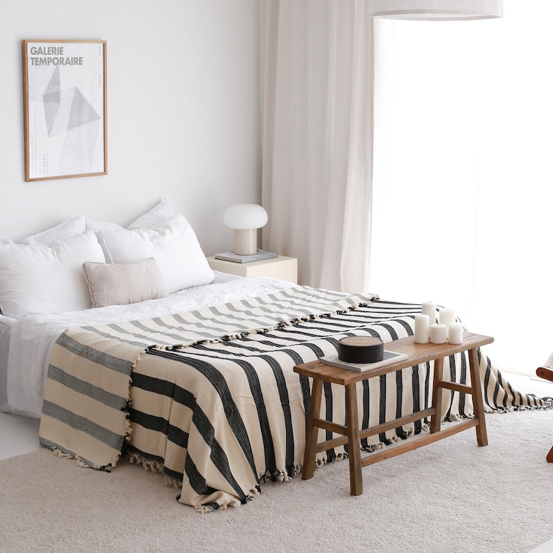 Op de afbeelding: Een zwart-wit gestreepte deken gedrapeerd over een bed met witte lakens. Het bed staat in een slaapkamer met een witte muur en een raam met witte gordijnen. Een houten kruk staat voor het bed.