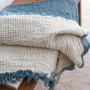 May include: A soft, white throw blanket with a blue fringe. The blanket is folded and draped over a wooden bench.