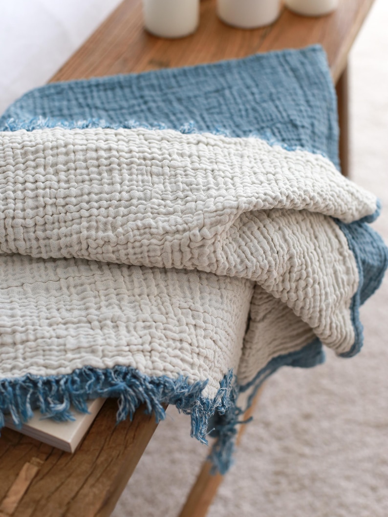 May include: A soft, white throw blanket with a blue fringe. The blanket is folded and draped over a wooden bench.