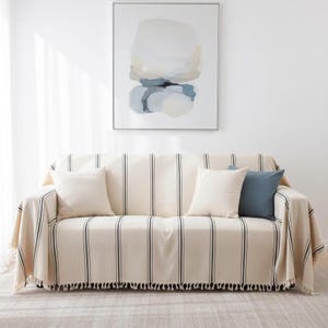 Puede incluir: Un sofá color crema está cubierto con una manta a rayas con líneas verticales negras y un borde de flecos. Tres cojines cuadrados color crema y un cojín azul descansan sobre el sofá. Una pintura abstracta cuelga de la pared sobre el sofá.