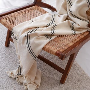 May include: A cream-colored throw blanket with black stripes draped over a wooden chair with a woven seat and back. The blanket has a fringed edge and a textured, woven pattern. The chair is on a light-colored rug.