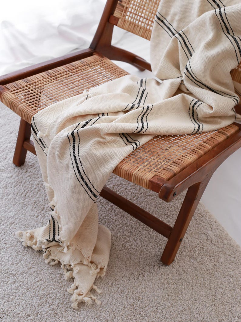May include: A cream-colored throw blanket with black stripes draped over a wooden chair with a woven seat and back. The blanket has a fringed edge and a textured, woven pattern. The chair is on a light-colored rug.