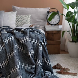 Peut inclure: Une couverture à rayures bleu marine et blanches avec une bordure à franges est drapée sur un lit. La couverture a un aspect tissé texturé. Le lit est fait avec des oreillers gris et blancs. Un réveil noir et une plante en pot sont en arrière-plan.