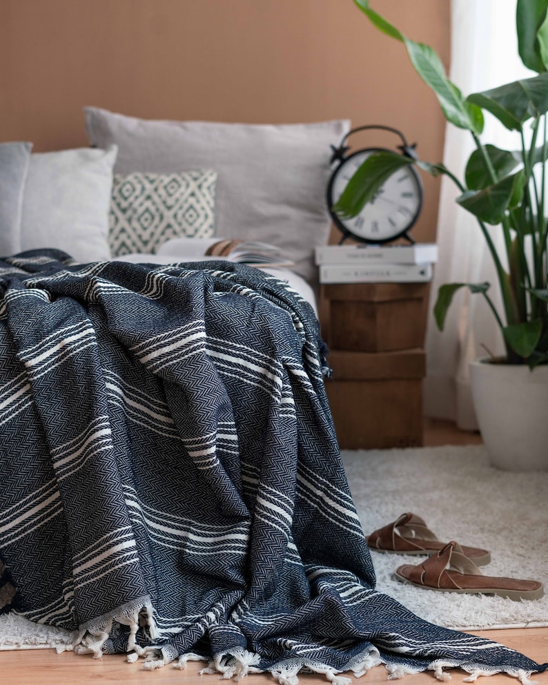 May include: A navy blue and white striped blanket with a fringed edge is draped over a bed. The blanket has a textured, woven appearance. The bed is made up with pillows and a neutral color scheme. A black alarm clock and a potted plant are visible in the background.