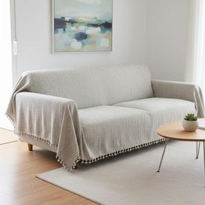 May include: A light gray sofa blanket with a textured weave and a fringe of small black pom-poms. The blanket is draped over a light gray sofa. A painting hangs on the wall above the sofa. A small round table and rug are in front of the sofa.