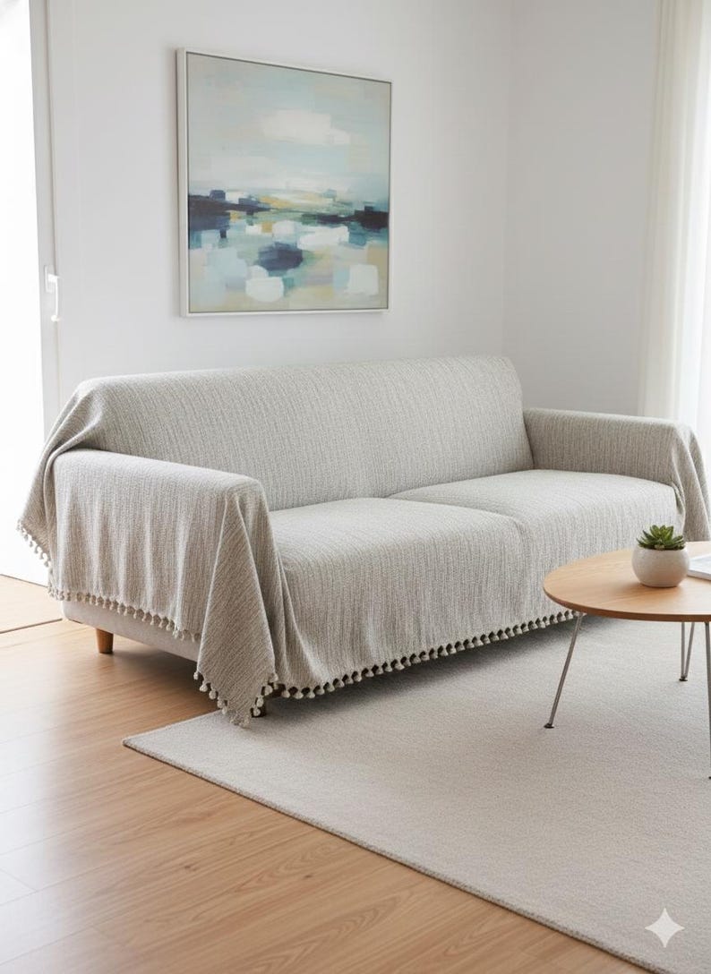 May include: A light gray sofa blanket with a textured weave and a fringe of small black pom-poms. The blanket is draped over a light gray sofa. A painting hangs on the wall above the sofa. A small round table and rug are in front of the sofa.