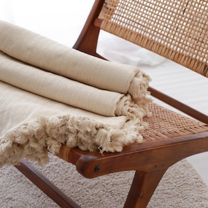 Op de afbeelding: Een beige deken met franjes is gevouwen en geplaatst op een houten stoel met een geweven rotan zitting.