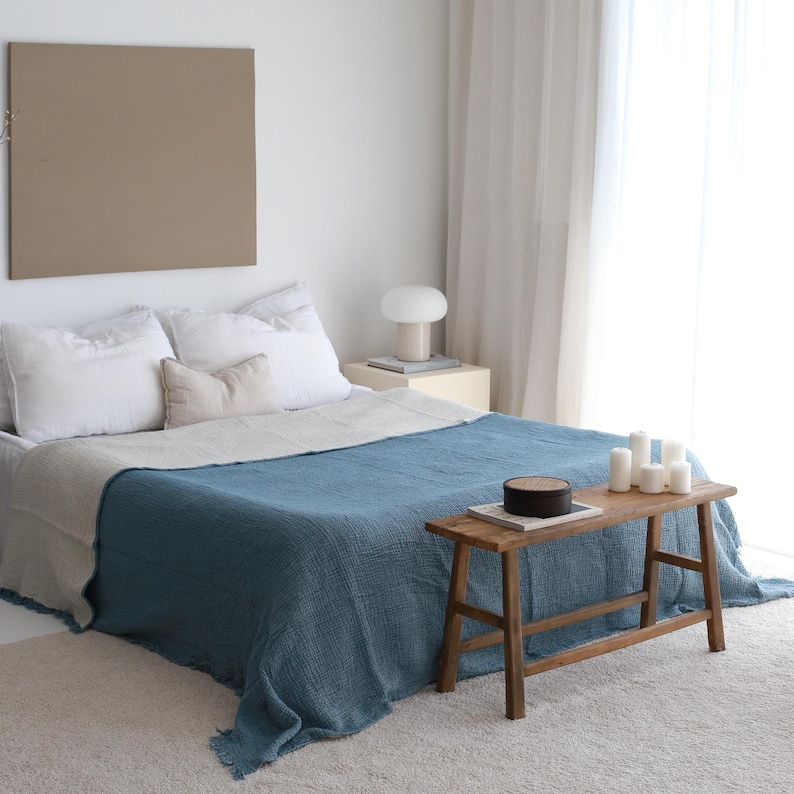 May include: A blue and white throw blanket draped over a bed with white pillows. A wooden bench with a round box on top sits in front of the bed.