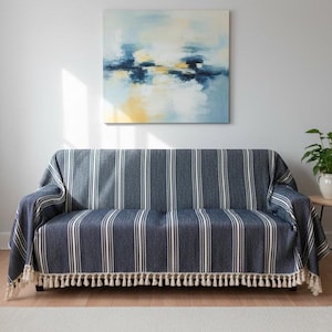 May include: A navy blue and white striped sofa cover with a cream-coloured tassel fringe. The cover is draped over a sofa, with a painting on the wall above. The painting has blue, yellow, and white colours.