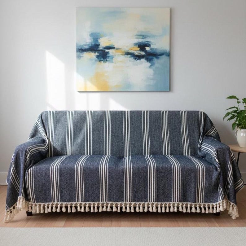 May include: A navy blue and white striped sofa cover with a cream-colored tassel fringe. The cover is draped over a sofa, with a painting on the wall above. The painting has blue, yellow, and white colors.