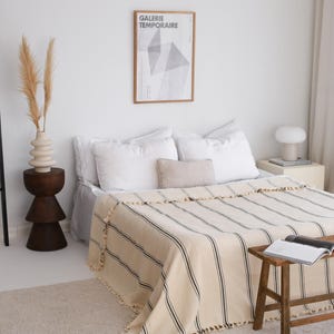May include: A bedroom scene featuring a bed with white linens and a striped blanket in cream and black. A wooden stool holds a book. A framed poster with the text "GALERIE TEMPORAIRE" hangs on the wall. A vase with dried plants sits on a wooden side table.