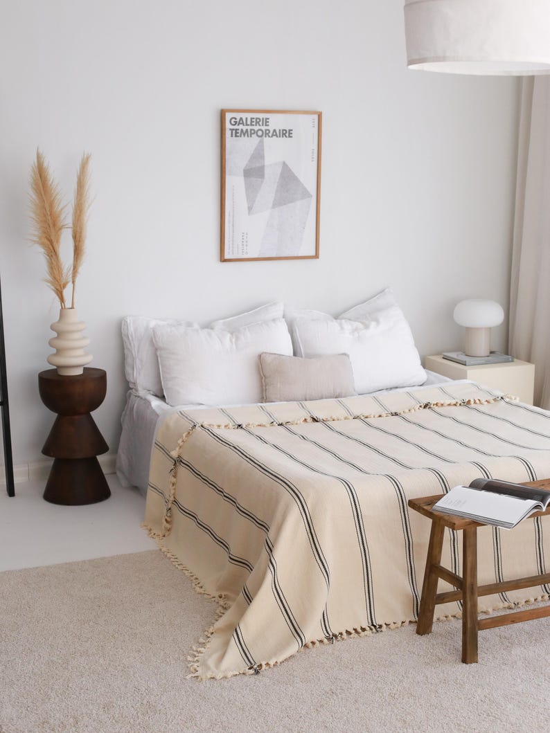 May include: A bedroom scene featuring a bed with white linens and a striped blanket in cream and black. A wooden stool holds a book. A framed poster with the text "GALERIE TEMPORAIRE" hangs on the wall. A vase with dried plants sits on a wooden side table.