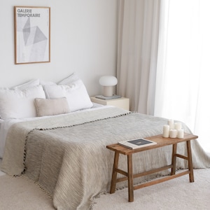 Op de afbeelding: Een wit bed met een grijs en wit wollen deken en een houten bank met kaarsen erop. Het bed staat in een witte kamer met witte gordijnen en een ingelijst poster met de tekst "Galerie Temporaire".