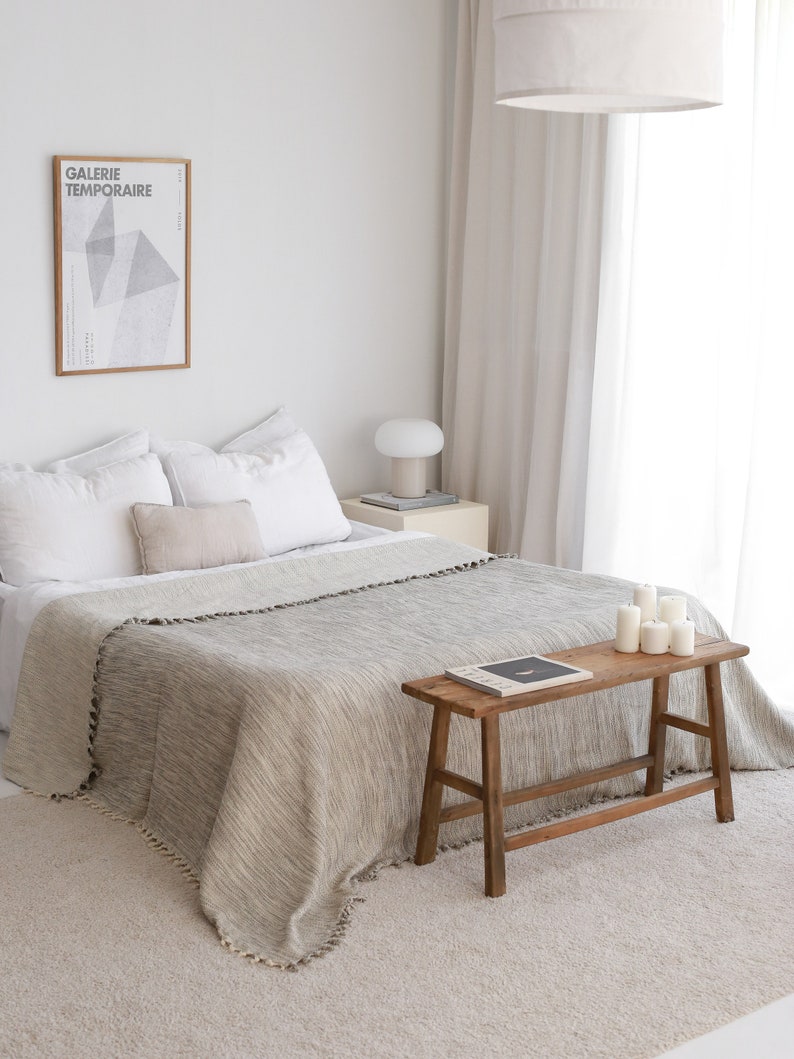 Op de afbeelding: Een wit bed met een grijs en wit wollen deken en een houten bank met kaarsen erop. Het bed staat in een witte kamer met witte gordijnen en een ingelijst poster met de tekst "Galerie Temporaire".