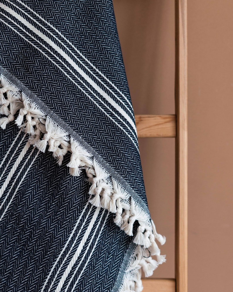May include: A navy blue and white striped throw blanket with a herringbone pattern. The blanket has white fringe along the edge. The blanket is draped over a wooden ladder.