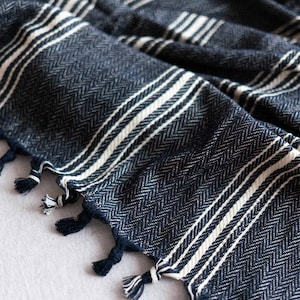 May include: Close-up of a navy blue and white striped woven textile. The fabric has a herringbone pattern and white stripes. The edges are finished with navy blue tassels. The textile is likely a blanket or throw.