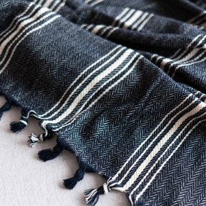 May include: Close-up of a navy blue and white striped woven textile. The fabric has a herringbone pattern and white stripes. The edges are finished with navy blue tassels. The textile is likely a blanket or throw.
