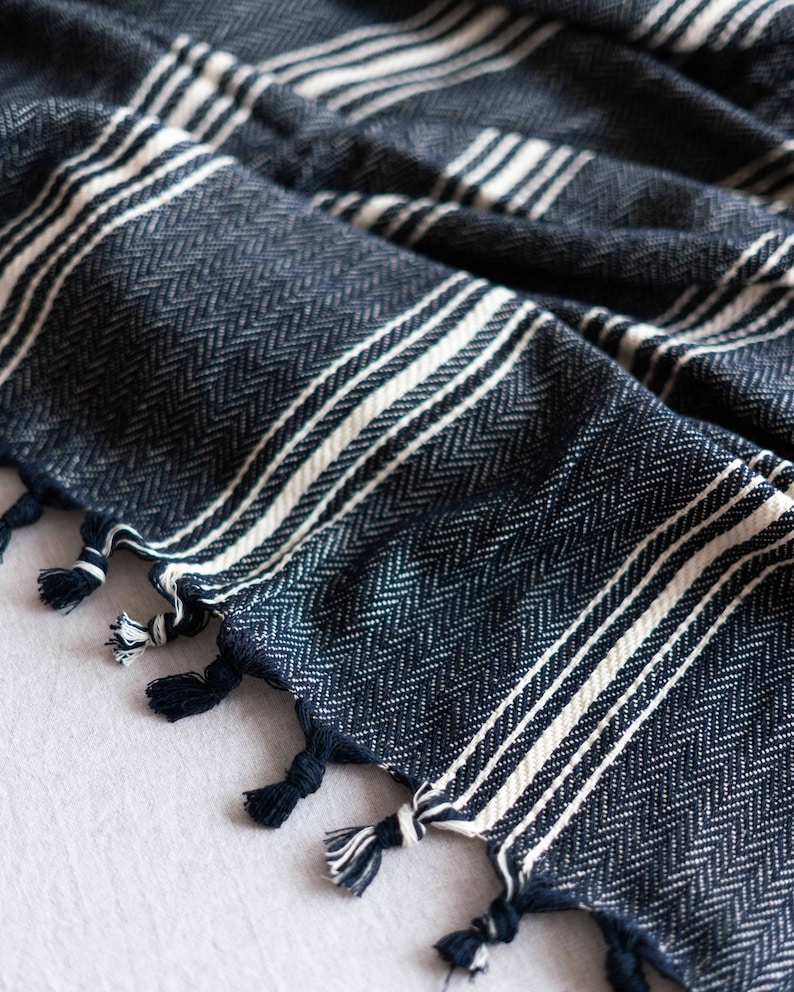 May include: Close-up of a navy blue and white striped woven textile. The fabric has a herringbone pattern and white stripes. The edges are finished with navy blue tassels. The textile is likely a blanket or throw.