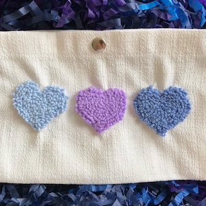 May include: White pouch with three embroidered hearts. The hearts are blue, purple, and light blue.