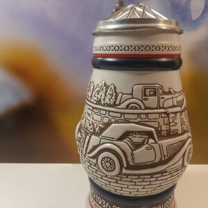 K&ouml;nnte beinhalten: Ein wei&szlig;er Keramik-Bierkrug mit einem silbernen Deckel und einem rot-wei&szlig;-blauen Band um den oberen Rand. Der Krug zeigt eine detaillierte Illustration von zwei Oldtimern auf einer gepflasterten Stra&szlig;e.