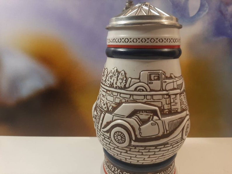 K&ouml;nnte beinhalten: Ein wei&szlig;er Keramik-Bierkrug mit einem silbernen Deckel und einem rot-wei&szlig;-blauen Band um den oberen Rand. Der Krug zeigt eine detaillierte Illustration von zwei Oldtimern auf einer gepflasterten Stra&szlig;e.
