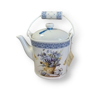 May include: White ceramic teapot with a blue and white floral design. The teapot has a metal handle and a lid with a blue ribbon. The teapot is sitting on a white surface.