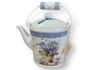 May include: White ceramic teapot with a blue and white floral design. The teapot has a metal handle and a lid with a blue ribbon. The teapot is sitting on a white surface.