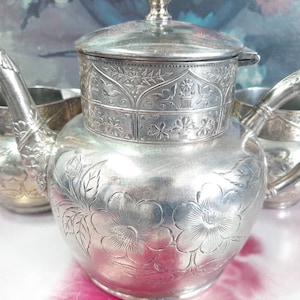 May include: A silver teapot with an ornate floral design and a lid. The teapot has a bird and floral motif on the lid and a floral design on the body. The teapot is part of a tea set.